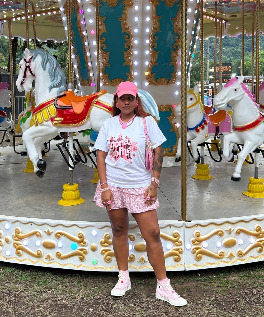 Pessoa vestindo camiseta, saia florida e tênis All Star rosa, em frente a carrossel com cavalos.