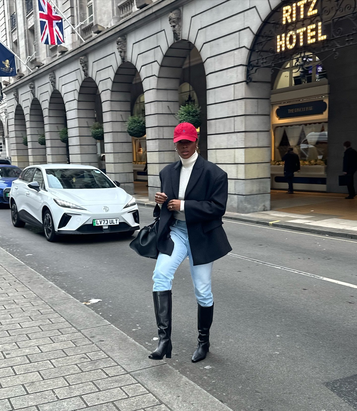 Pessoa com calça jeans, blazer preto e boné vermelho caminha em frente ao Ritz Hotel.