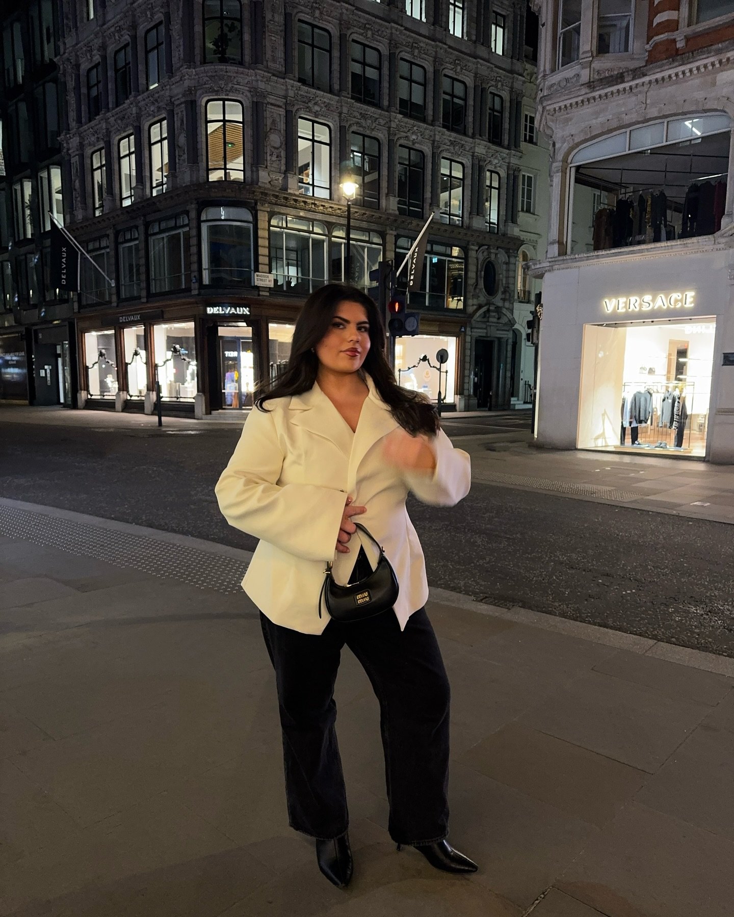 A pessoa veste um blazer branco e segura uma bolsa preta, na frente de lojas iluminadas em uma rua à noite.