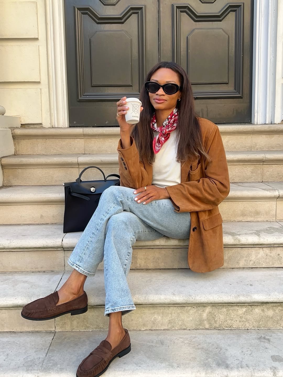 Pessoa sentada em degraus, vestindo calça jeans e blazer marrom, segurando café, com bolsa preta ao lado.