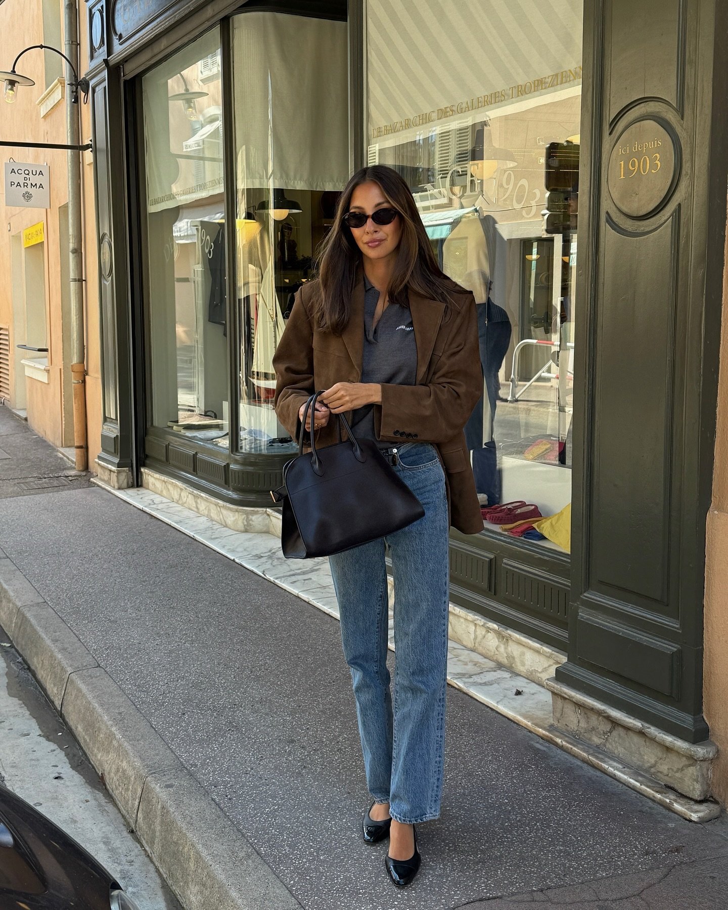Pessoa na calçada usando calça jeans e blazer marrom, carregando bolsa preta e usando óculos escuros. Fundo com vitrine de loja.