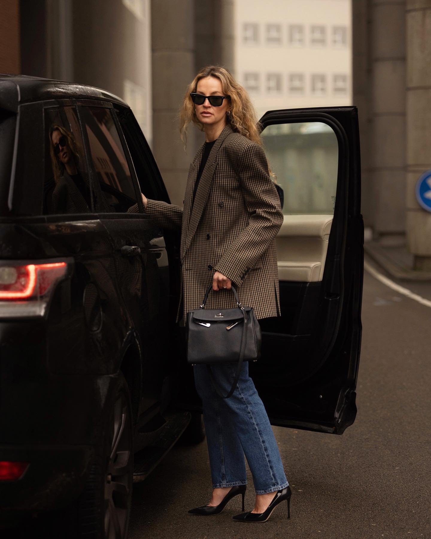 Pessoa em pé ao lado de carro, usando calça jeans e blazer xadrez, segurando uma bolsa preta.