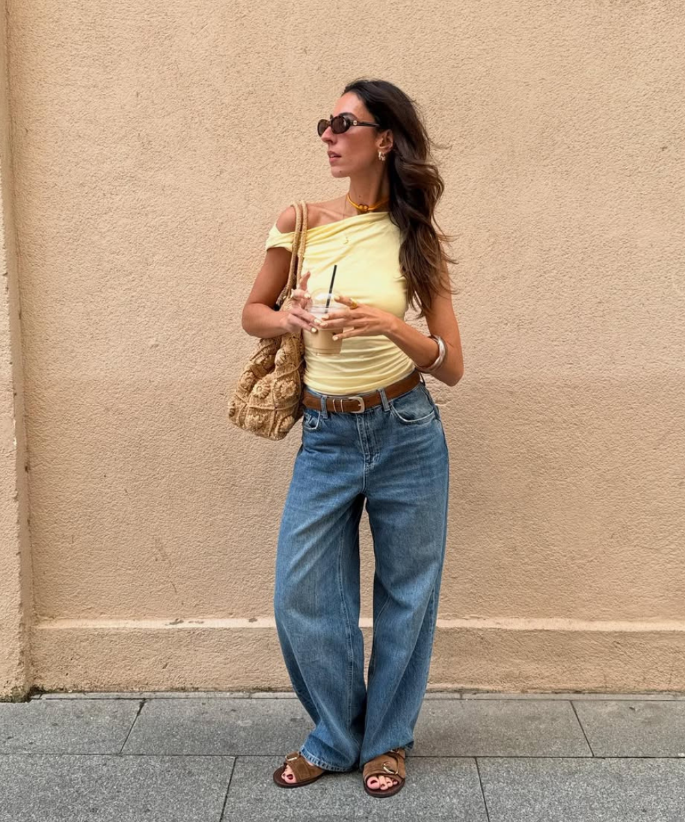 Mulher de blusa amarela e jeans largo segura bebida e usa papete marrom, encostada em parede creme.