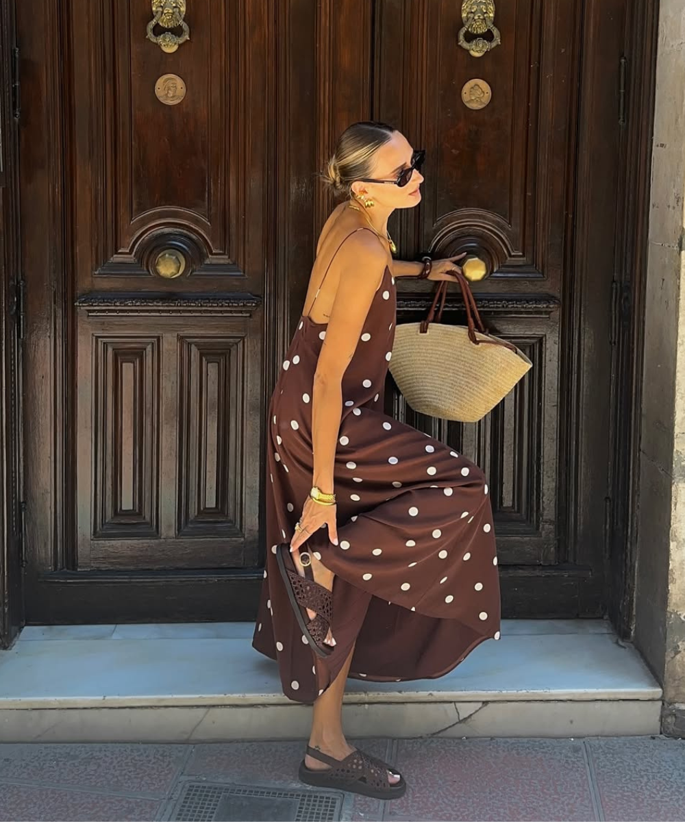 Mulher em vestido marrom de bolinhas, segurando papete marrom e bolsa de palha em frente a uma porta de madeira.