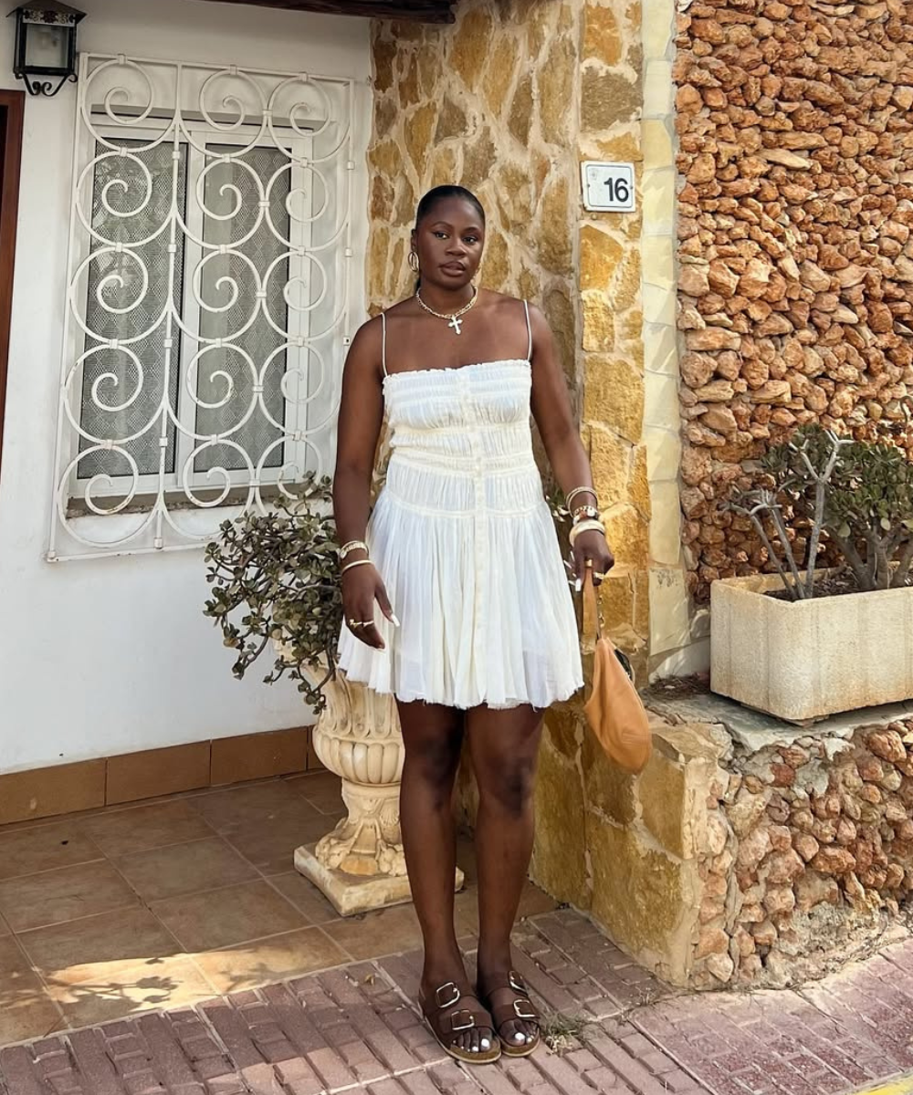 Mulher em vestido branco, segurando bolsa marrom, usa papete marrom em frente a parede de pedra e janela com grades.