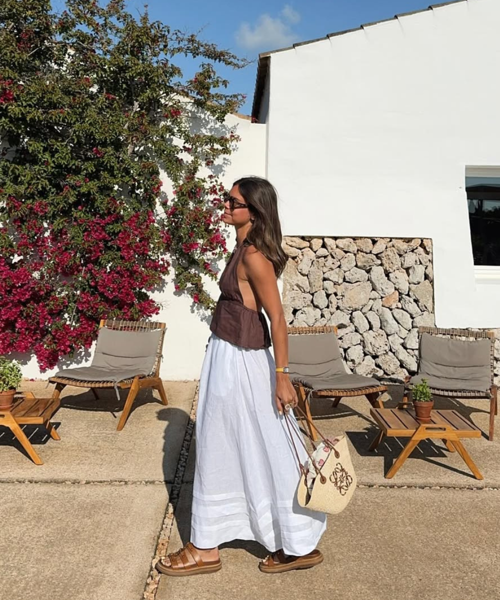 Pessoa com saia branca e blusa marrom, caminhando em varanda. Usa papete marrom e carrega bolsa bege.