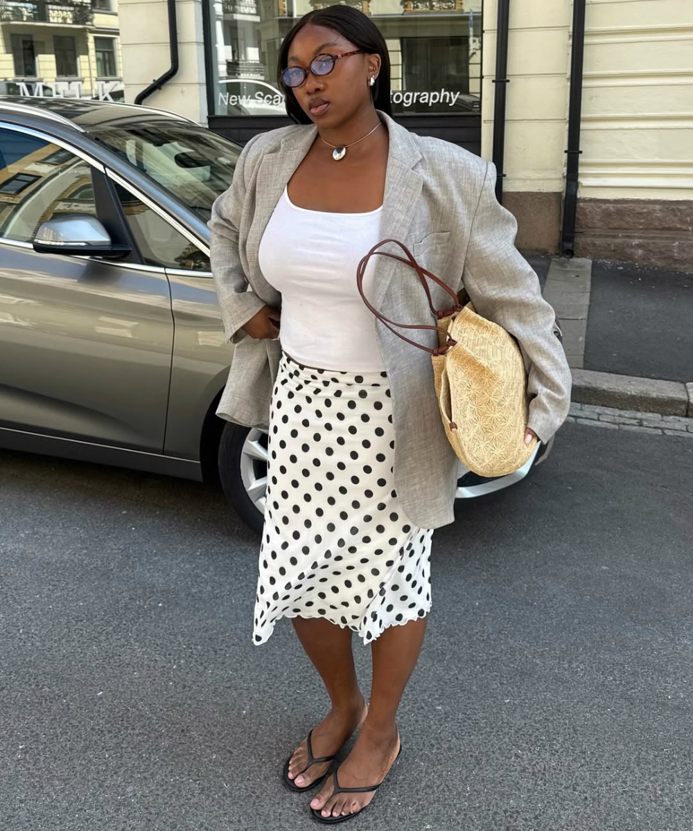 Pessoa vestindo saia branca de bolinhas, blusa branca, blazer cinza e sandálias, segurando bolsa beige.