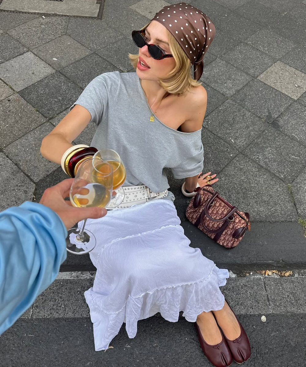 Uma pessoa com lenço marrom e saia branca está sentada na calçada, segurando um copo de bebida.