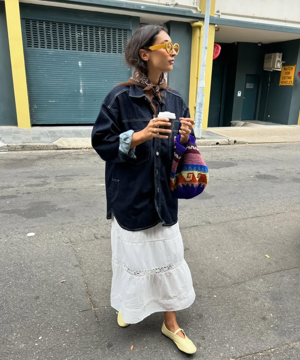 Mulher com saia e jaqueta jeans, segurando café. A bolsa colorida e óculos amarelos compõem o look casual. Inspiração de looks com saia branca.