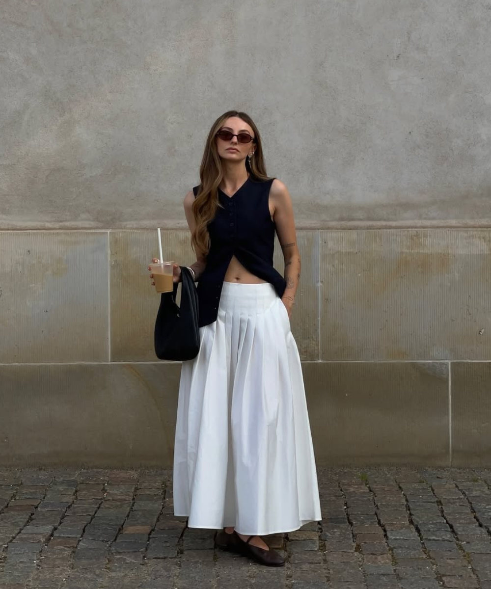 Pessoa com blusa preta e saia branca esvoaçante, óculos de sol e bolsa preta, segurando bebida.