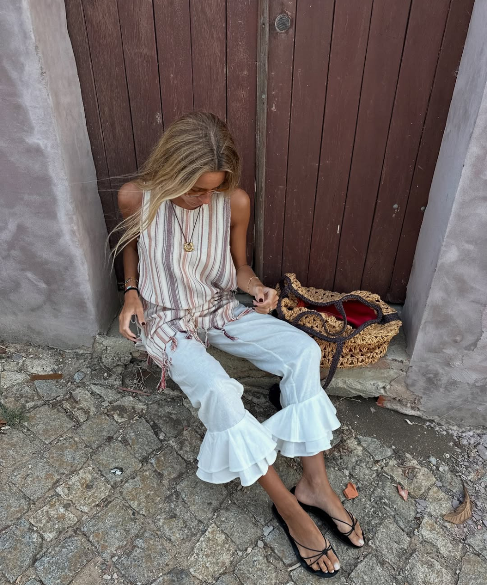 Pessoa sentada em calça branca com babados e sandália flat preta. Bolsa de palha ao lado.