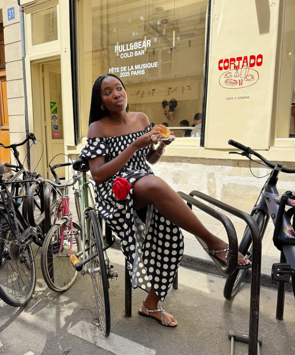 Pessoa com vestido de bolinhas pretas e brancas e sandália flat, segurando um croissant. Fundo com bicicletas e vitrine de café. Looks com sandália flat.