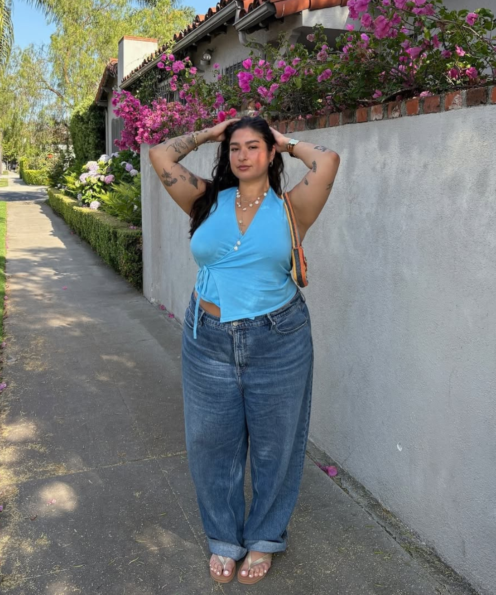 Pessoa em look casual com sandália flat, blusa azul e calça jeans solta, em uma calçada com flores.