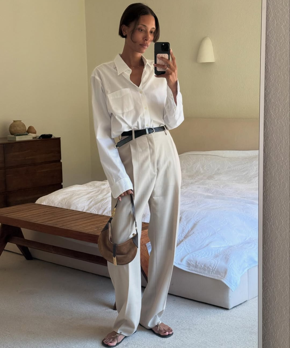 Pessoa em look casual com camisa branca, calça bege, cinto preto e sandálias flat, segurando bolsa marrom.