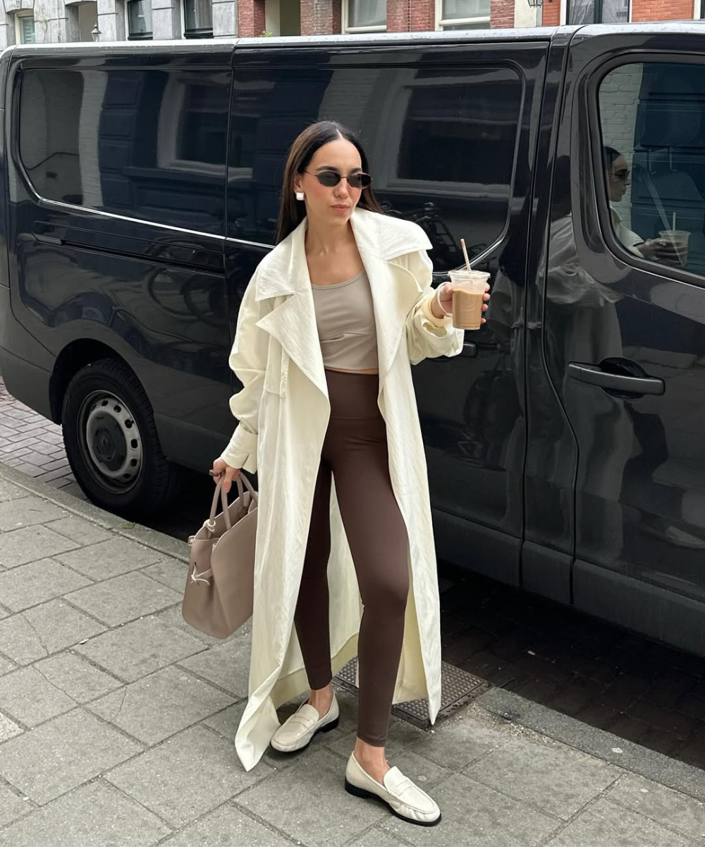 Mulher com calça marrom e casaco longo, segurando bebida e bolsa, passa por van preta. Look casual e confortável.