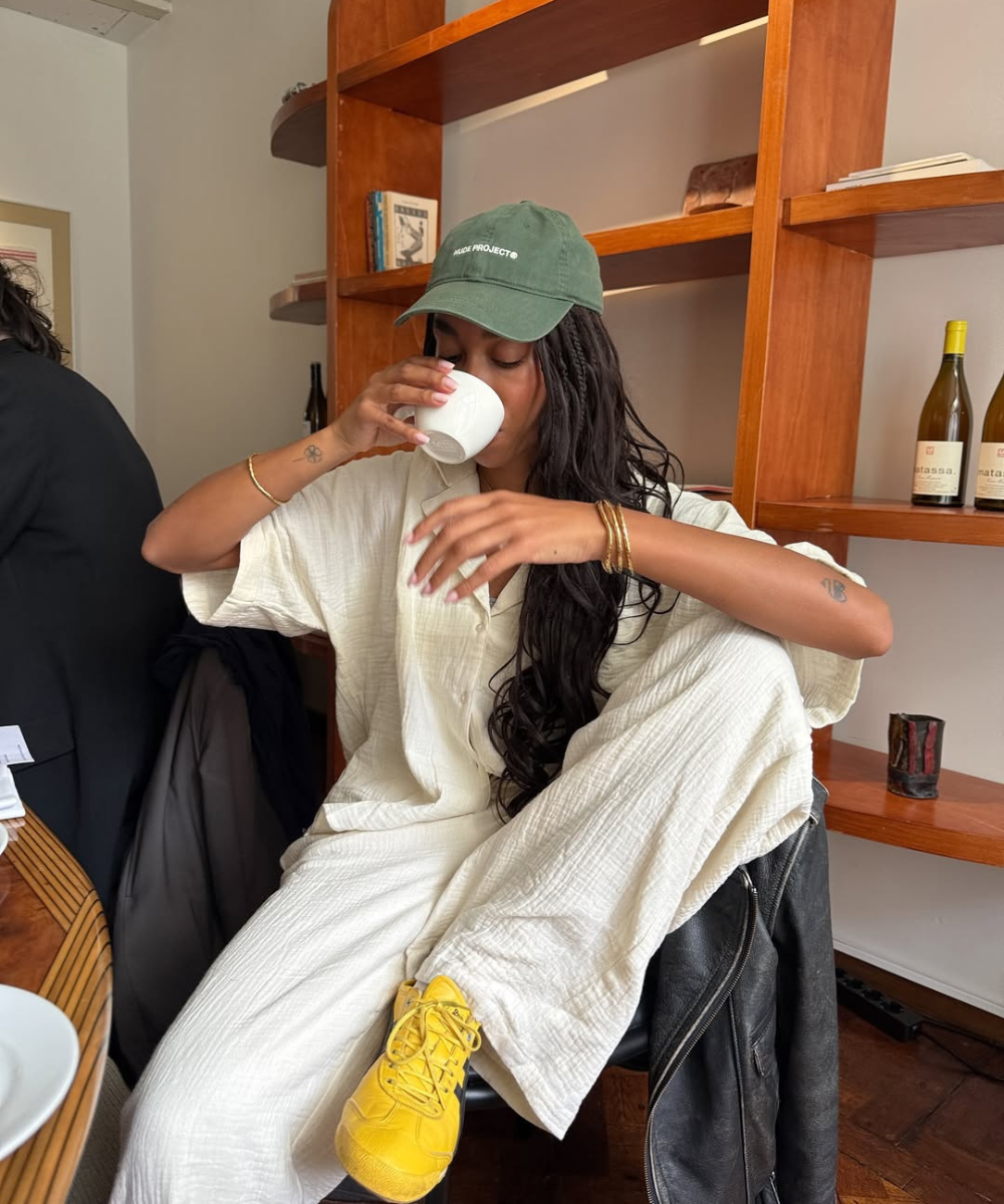 Pessoa relaxando, usando roupas confortáveis com calça clara e tênis amarelo, bebendo uma bebida em um ambiente casual.