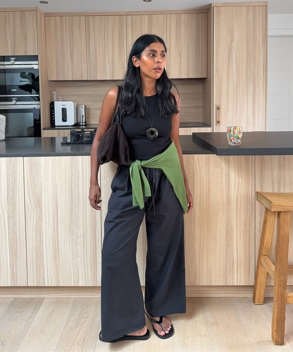 Pessoa com camisa preta e calça larga preta, blusa verde amarrada na cintura, em cozinha de madeira.