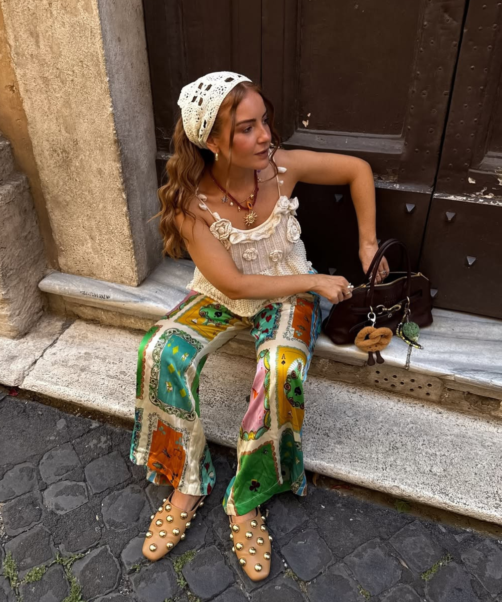 Mulher sentada em degrau, usando calça colorida estampada, sandálias com tachas e blusa de crochê.