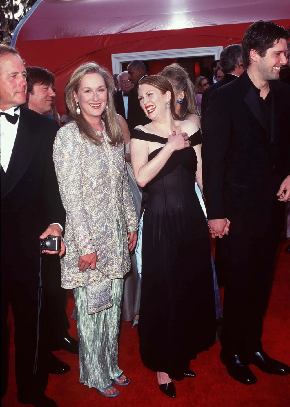 Celebridades em um evento de gala, Meryl Streep usa traje elegante bordado, sorrindo ao lado de amigos no tapete vermelho.