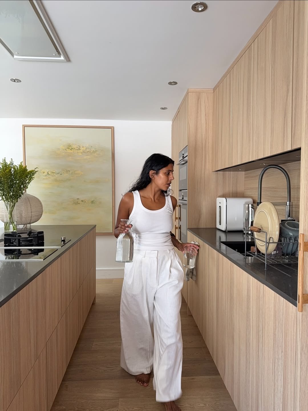Uma mulher em uma cozinha moderna, vestindo roupa branca, segurando uma garrafa e um copo. Look ano novo 2026