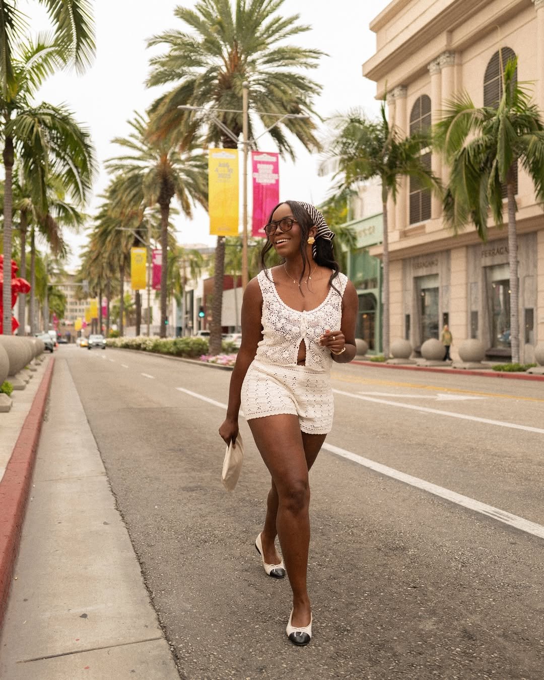 Mulher sorrindo caminha em rua arborizada, vestindo conjunto branco rendado. Looks modernos para o ano novo 2026.