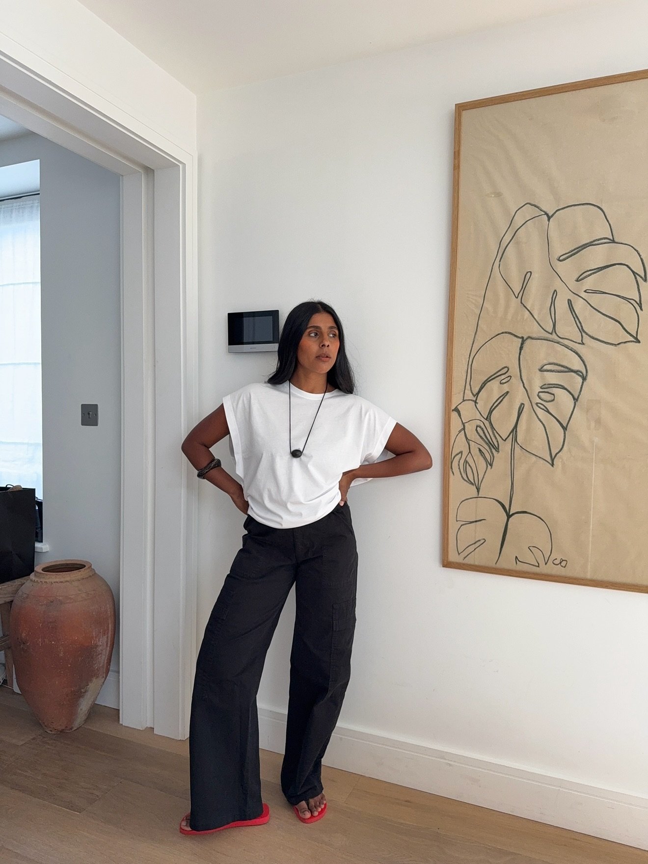 Pessoa em look casual com camiseta branca e calça preta ampla, ideal para primavera-verão. Pôster de folhagem ao fundo.