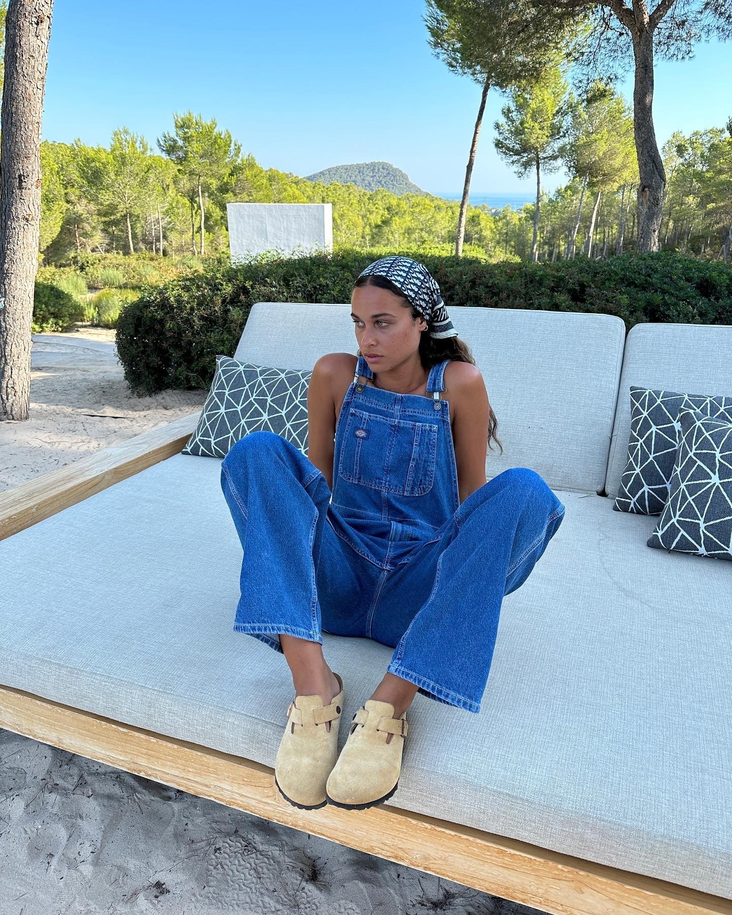 Pessoa com macacão jeans e lenço na cabeça, sentada em um sofá ao ar livre em cenário de natureza. Looks de primavera-verão.