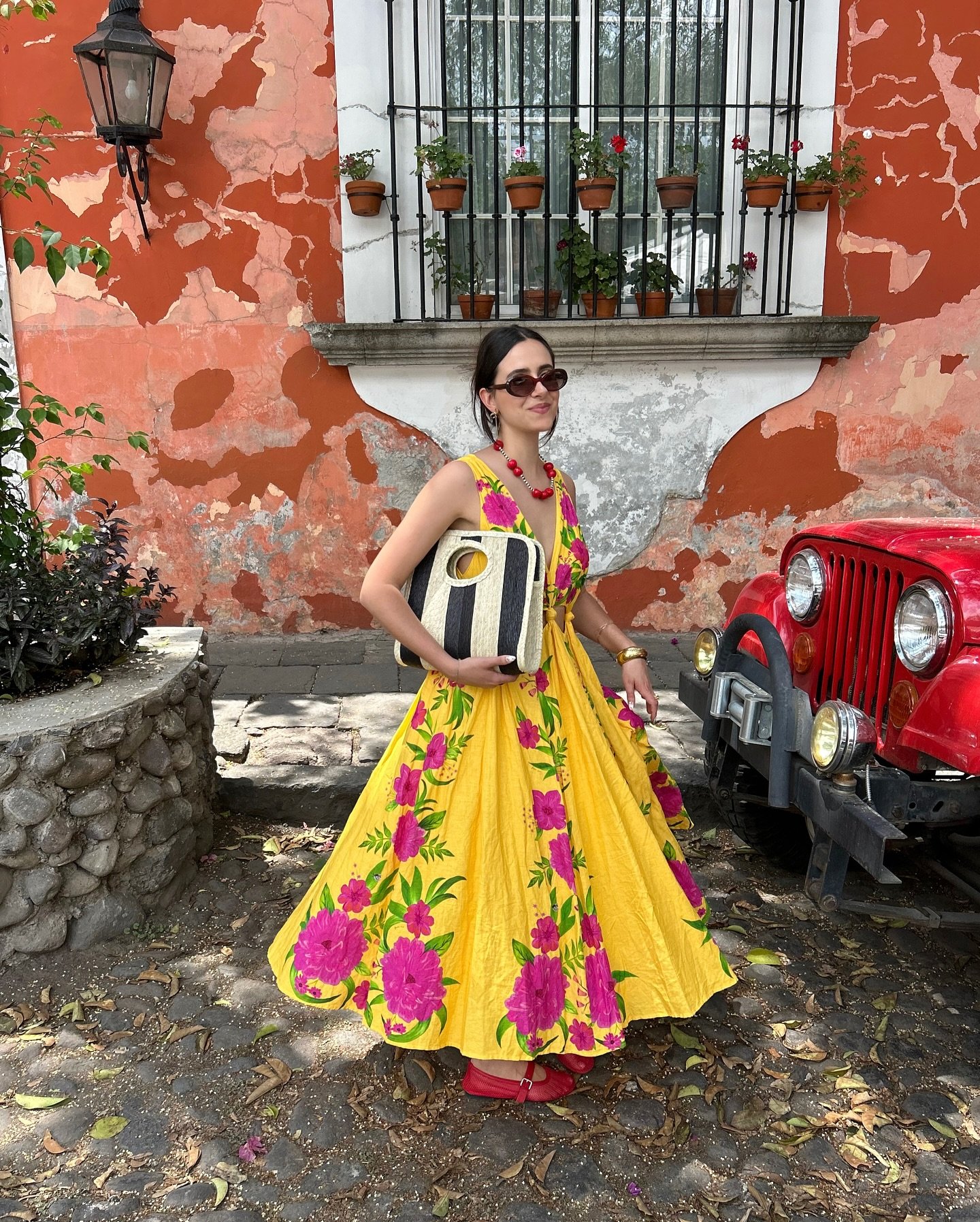 Mulher em vestido floral amarelo e rosa ao lado de jipe vermelho, em cenário com parede desgastada. Looks vibrantes primavera-verão.