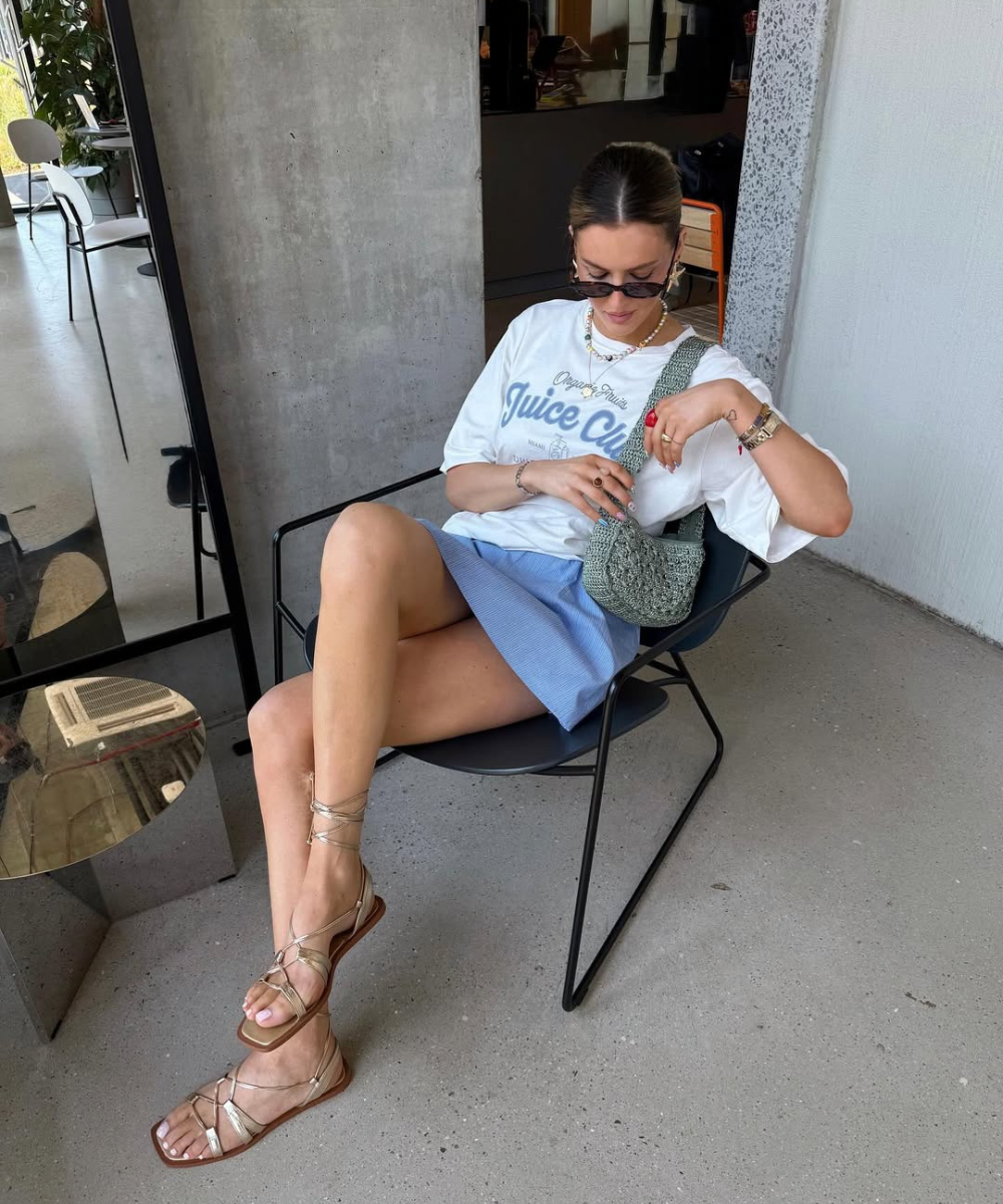 Mulher com camiseta branca e saia azul sentada, usando rasteirinha dourada, criando visuais bonitos e despojados.