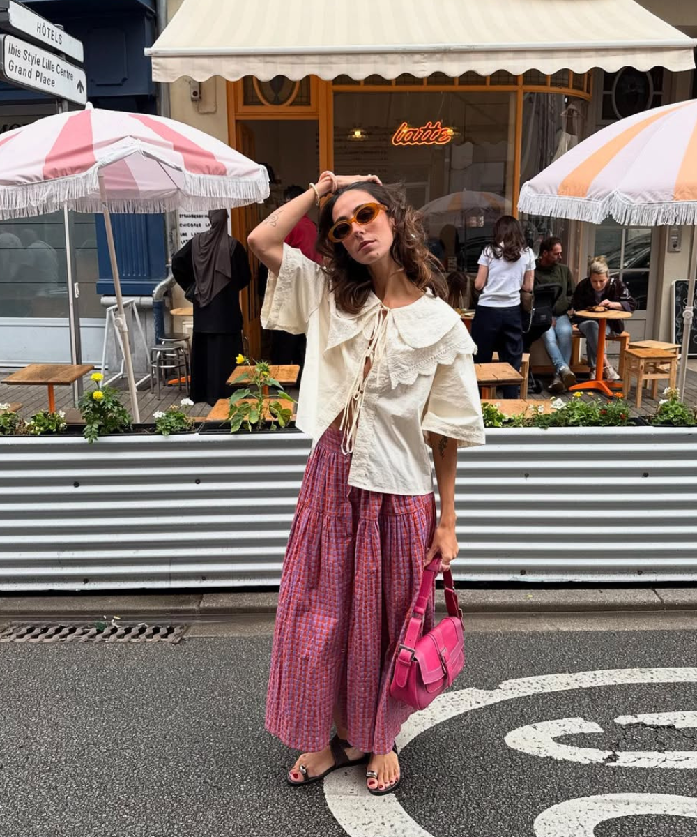 Pessoa com look estilo boho, vestindo blusa clara, saia longa, óculos escuros e rasteirinha, segura bolsa rosa.