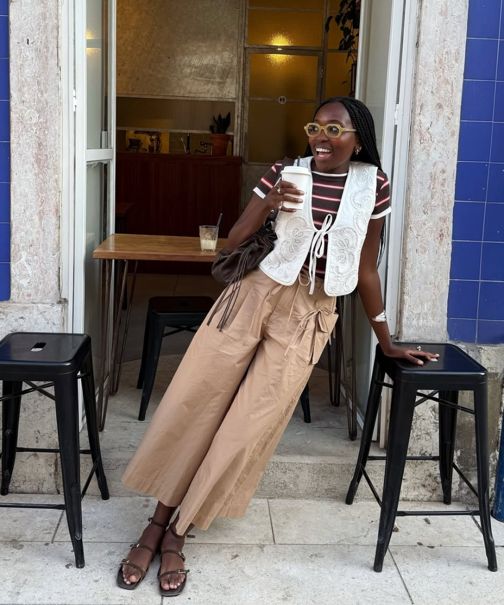 Pessoa sorrindo em frente a um café, vestindo calça bege, colete branco e rasteirinha marrom, segurando uma bebida.
