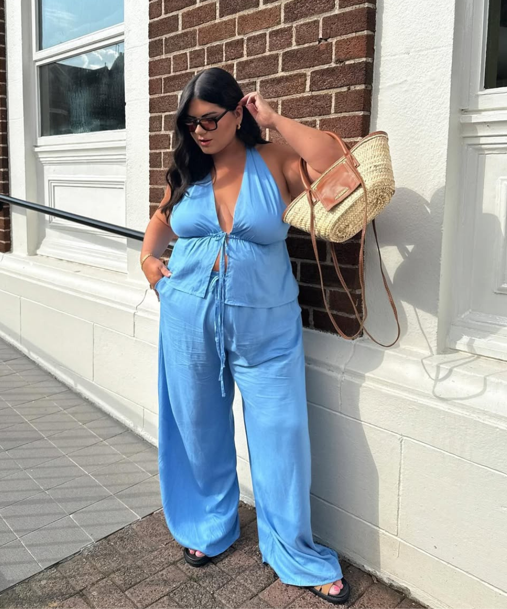 Mulher com conjunto azul e sandálias rasteiras pretas segura bolsa de palha, criando looks bonitos e casuais.