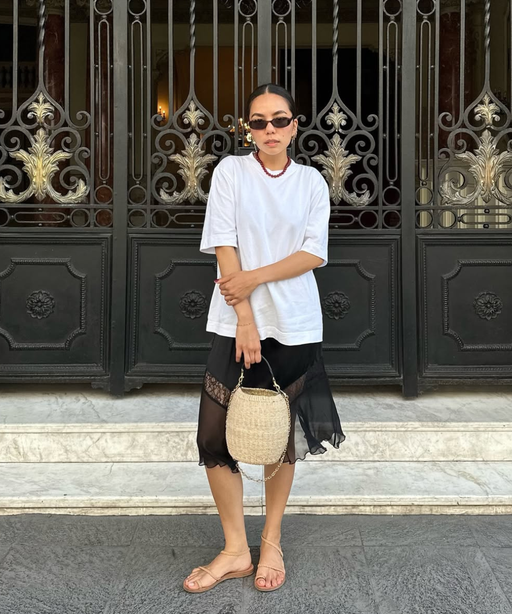 Mulher com camiseta branca, saia preta e rasteirinha, segurando bolsa de palha. Portão decorativo ao fundo.