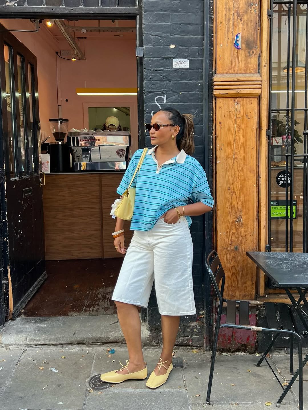 Mulher em frente a um café, usando camiseta listrada azul, bermuda branca e sapatilhas amarelas. Looks femininos estilosos.
