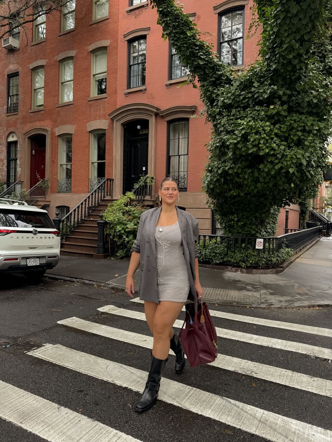 Mulher com looks femininos estilosos cruza a rua, vestindo blazer cinza, vestido claro e botas pretas, segurando uma bolsa vinho.