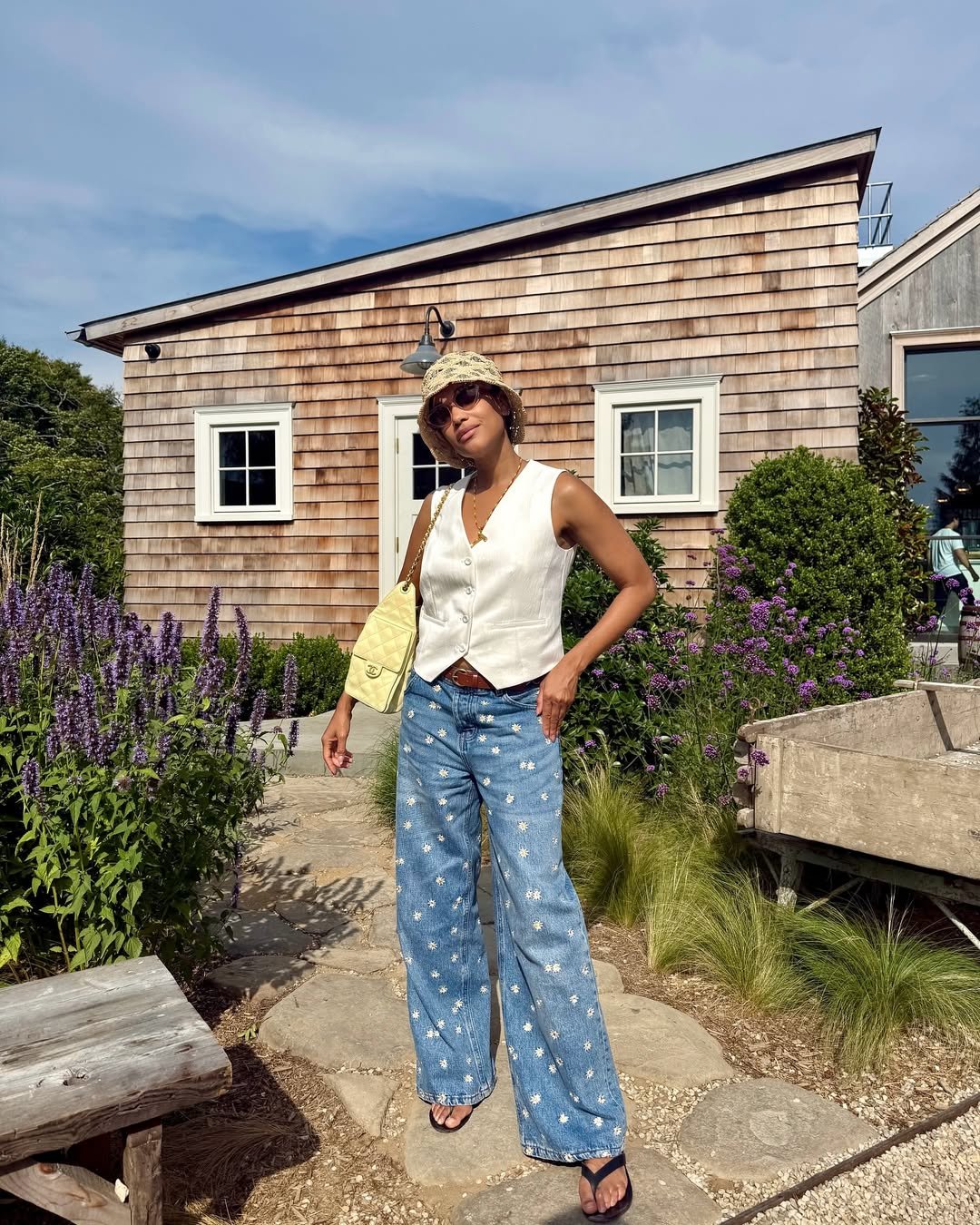 Mulher com colete branco e jeans florido posa frente a casa de madeira, combinando look estiloso com bolsa amarela e chapéu claro. Looks femininos estilosos