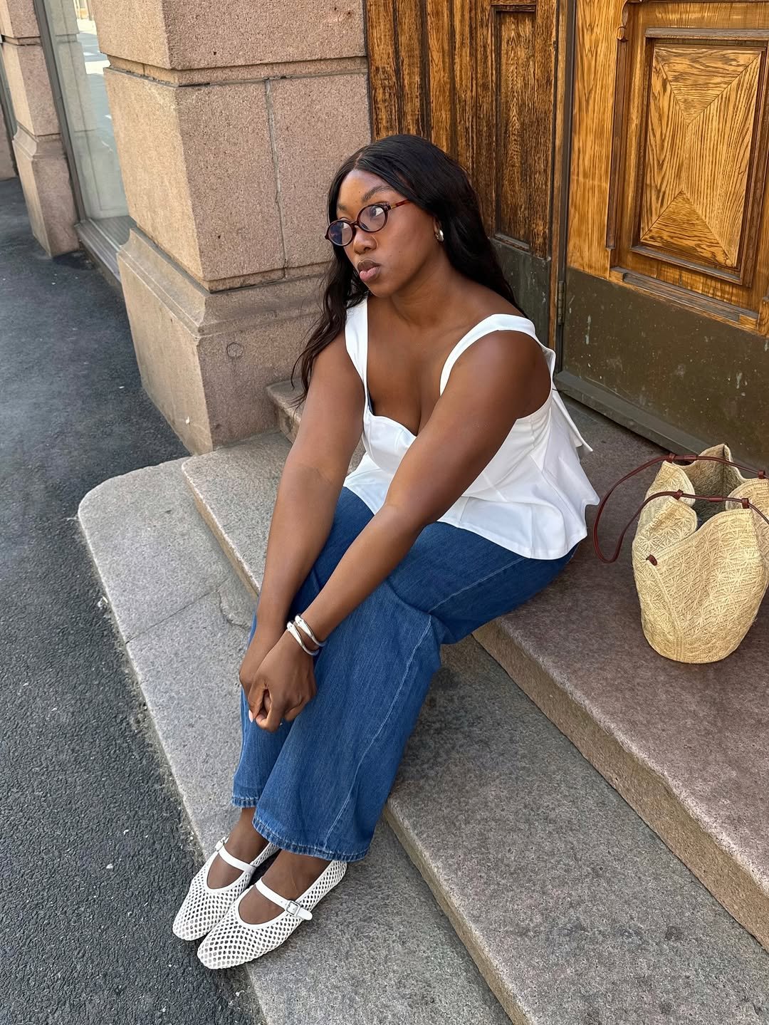 Mulher sentada em degraus, usando blusa branca e jeans, com saco de palha ao lado. Looks femininos estilosos