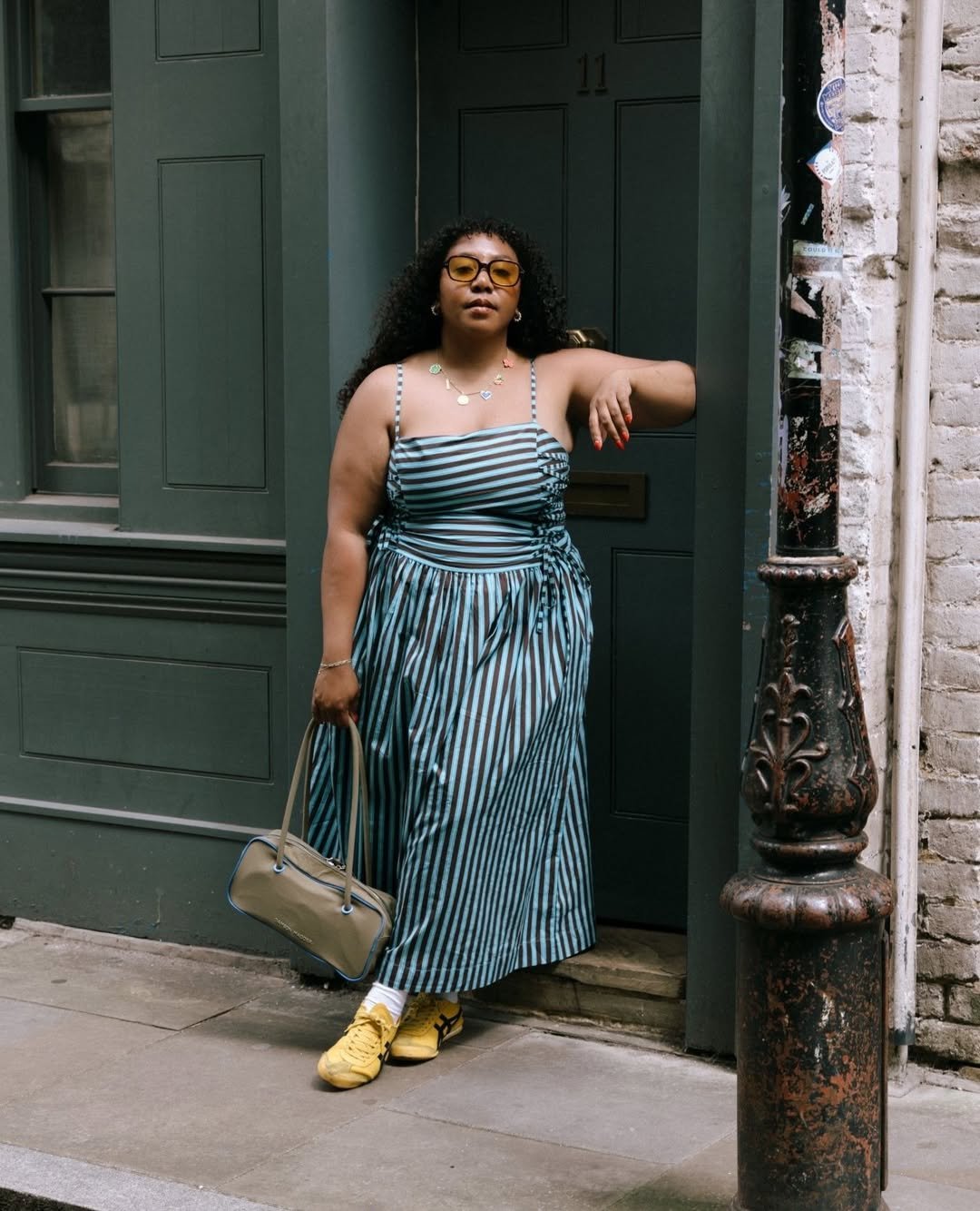 Vestido listrado azul e preto, mulher com óculos, bolsa bege e tênis amarelo, destacando looks femininos estilosos.