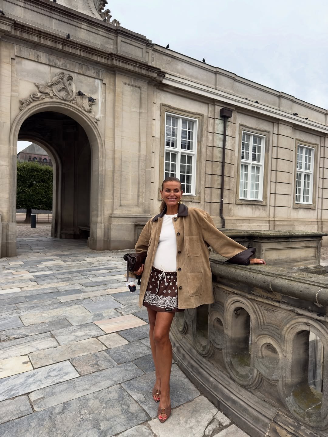 Mulher em look feminino e estiloso com casaco e saia estampada, em frente a um prédio histórico. Looks femininos estilosos