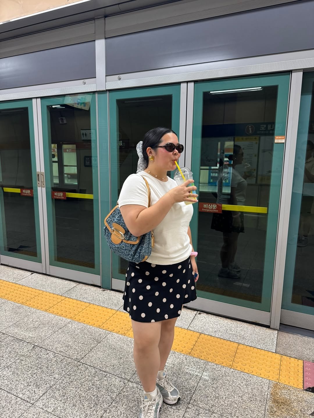 Mulher em estação de metrô com saia de bolinhas, camiseta branca e bolsa jeans, exemplificando looks femininos estilosos.