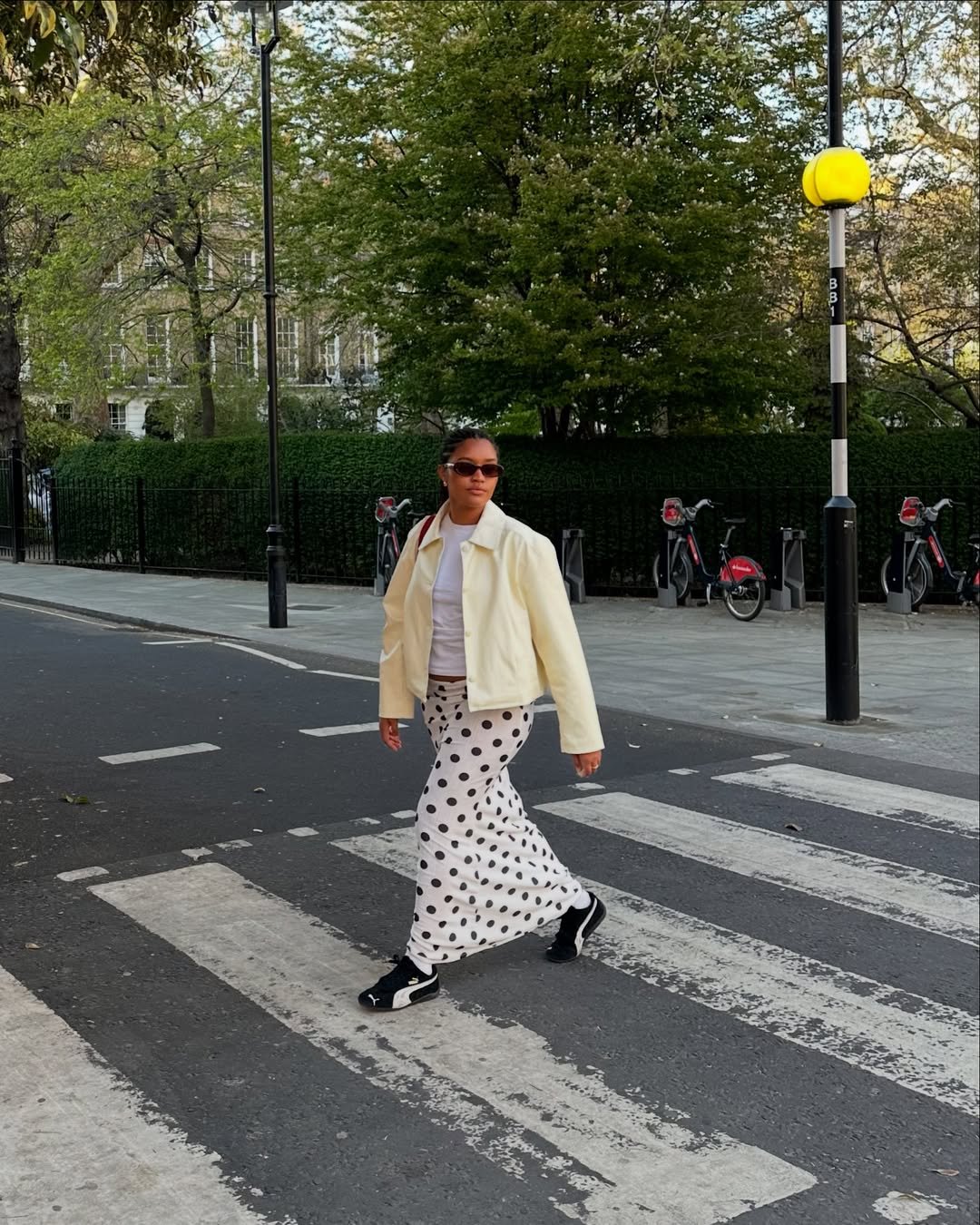 Pessoa atravessa faixa de pedestres com looks femininos estilosos, usando casaco claro e saia longa com bolinhas.