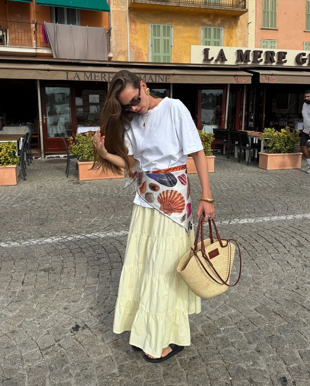 Mulher com saia longa amarela e blusa branca veste faixa estampada e segura bolsa de palha, criando looks femininos estilosos.