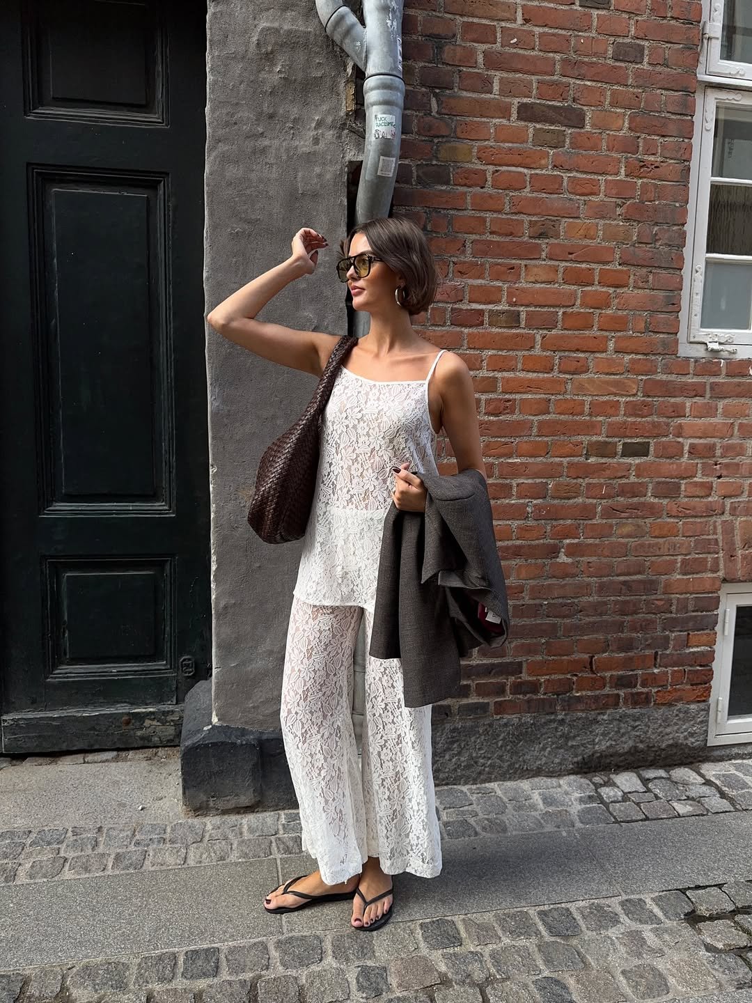 Pessoa com roupa branca de renda e chinelos em frente a parede de tijolos, carregando bolsa. inspiração para look de festival