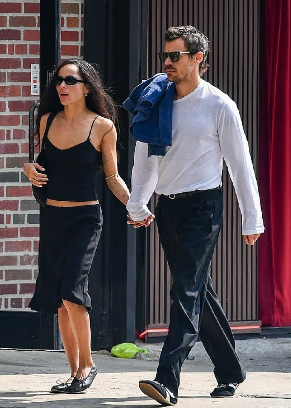 Zoë Kravitz e Harry Styles com roupas pretas e óculos de sol caminham de mãos dadas com outra de camisa branca, na calçada.