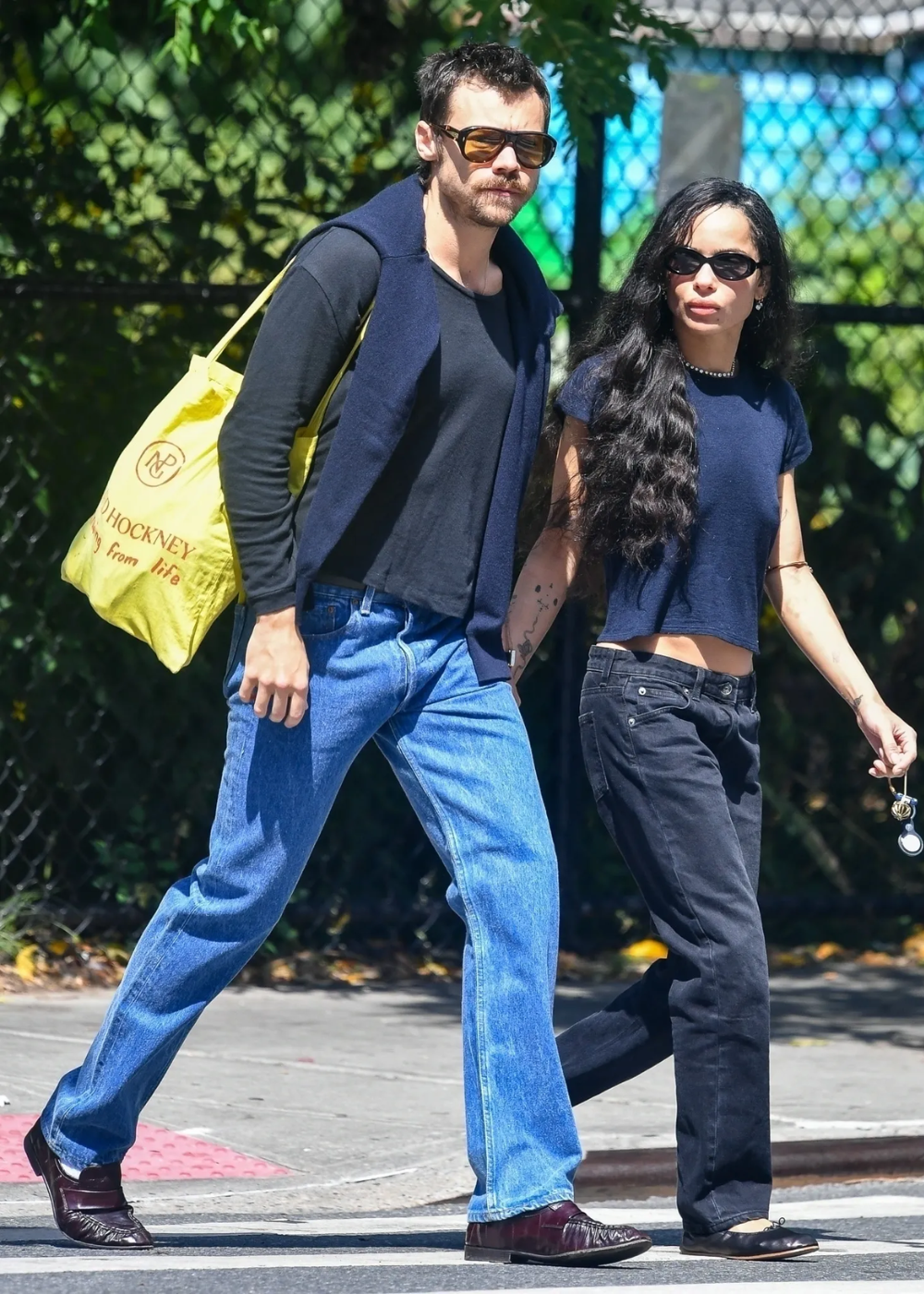 Zoë Kravitz e Harry Styles andando de mãos dadas em uma rua ensolarada, vestindo roupas casuais.