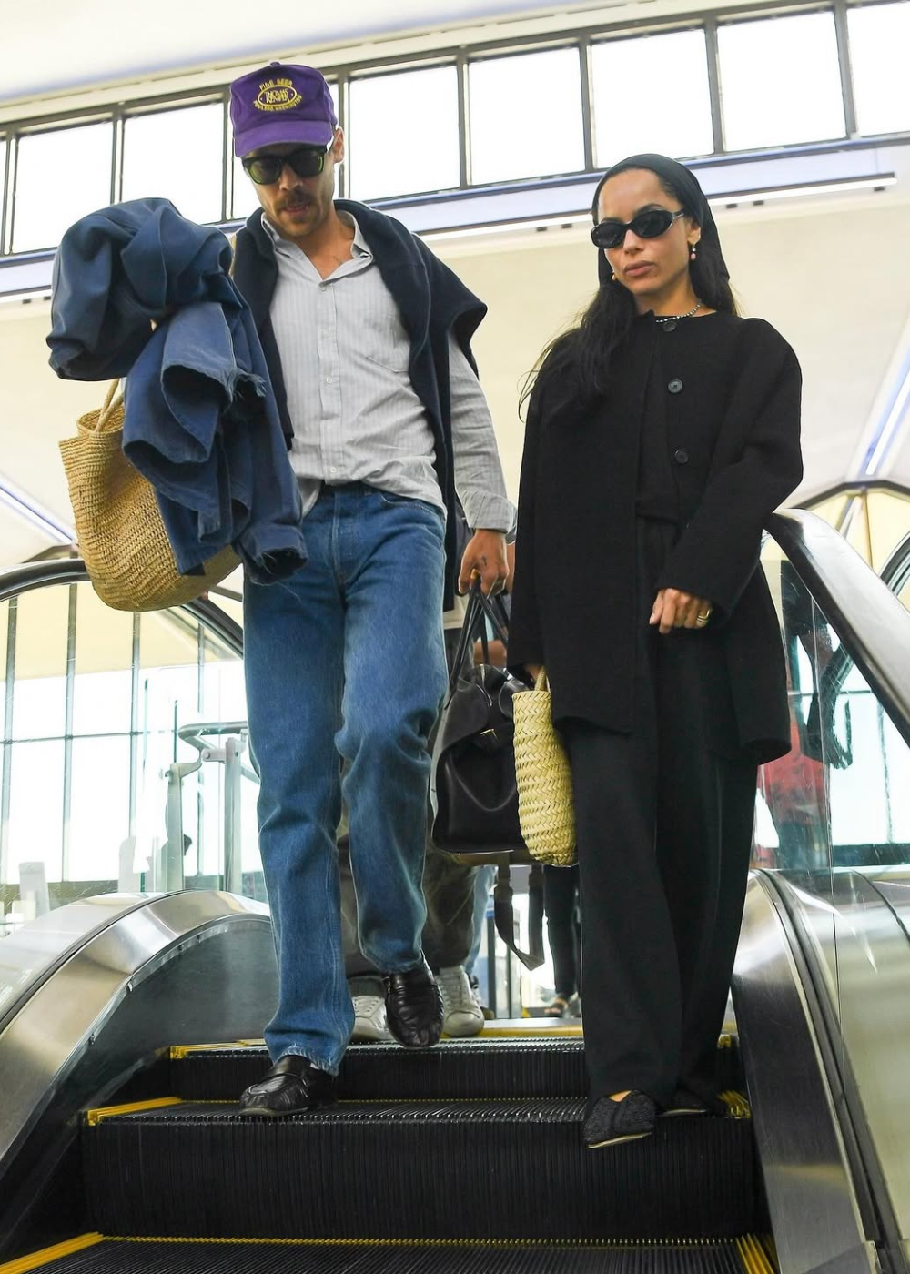 Zoë Kravitz e Harry Styles descem uma escada rolante, ambas usando óculos escuros e carregando bolsas.