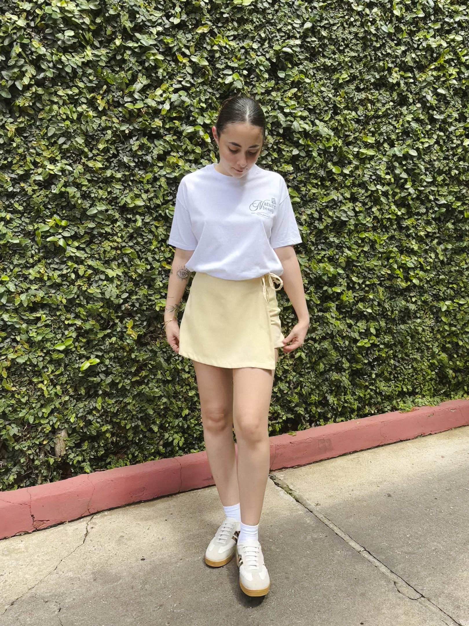 Pessoa com camiseta branca e saia amarela clara, em um fundo de folhagem verde, usando um dos looks de primavera estilosos.