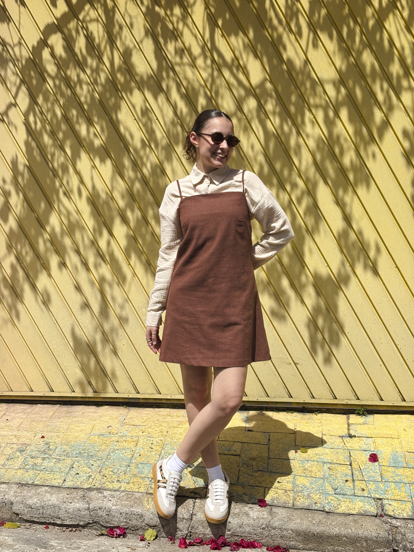 Pessoa usando vestido marrom sobre blusa clara, com tênis e óculos de sol. Fundo amarelo vibrante. Looks de primavera estilosos.