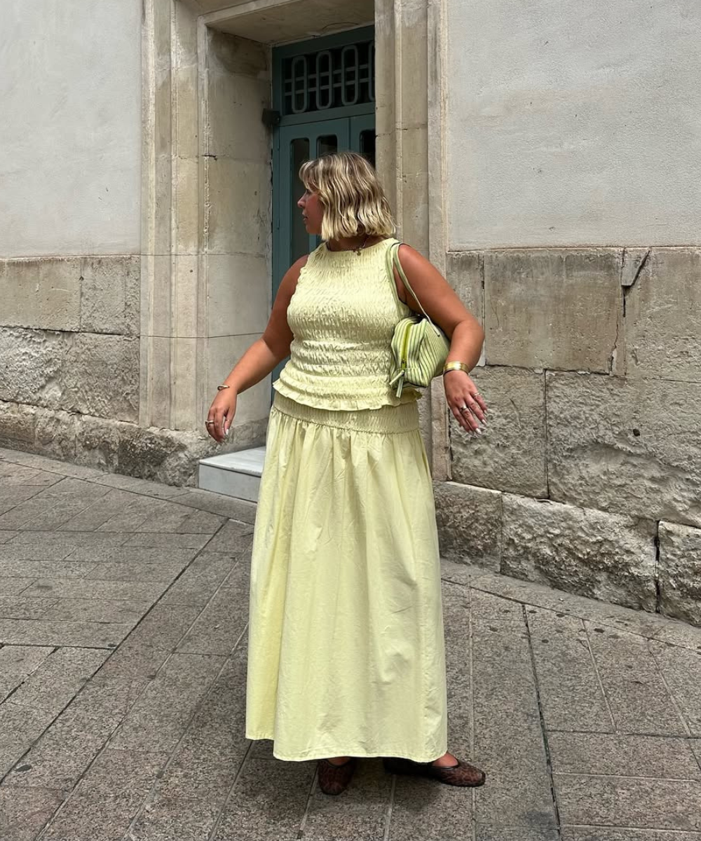 Mulher usa conjunto amarelo claro com saia longa, em frente à porta azul. Uma das tendências look para primavera.