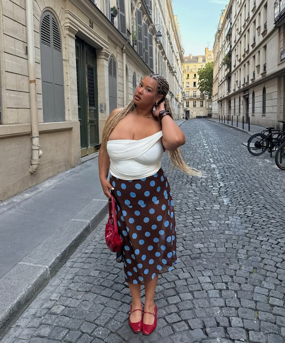Pessoa com saia de bolinhas azuis, top branco e bolsa vermelha, em rua de edifícios históricos. Looks tendência para primavera.